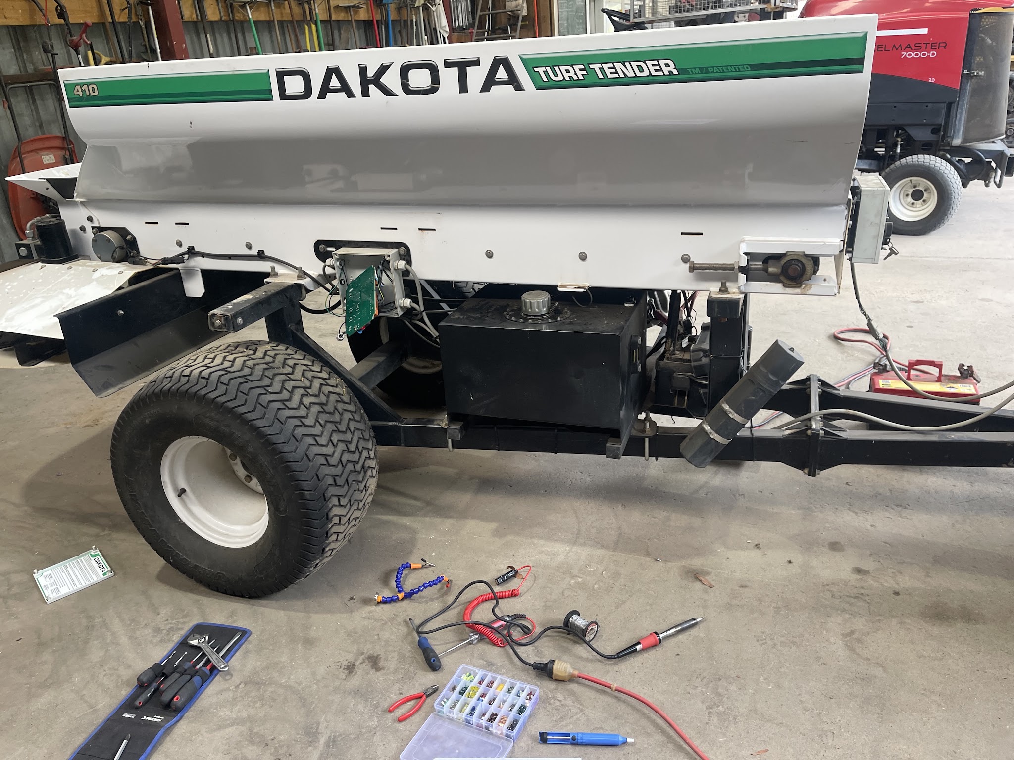 A Dakota turf tender with its control board exposed, alongside a Toro Reelmaster in the shed